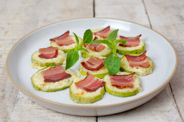 Homemade baked zucchini with cheese, crispy bacon and basil on a white wooden background served in a plate.
