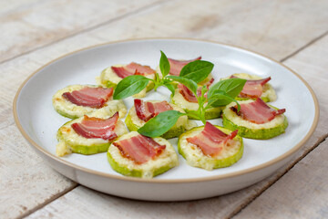 Homemade baked zucchini with cheese, crispy bacon and basil on a white wooden background served in a plate.	