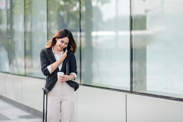 Young Asian businesswoman talking on phone and walking in airport before business trip. Beautiful woman passenger has mobile call and discusses something with smile, holds coffee in hand