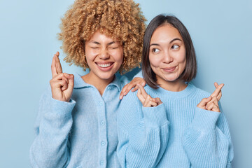 Luck and expectations concept. Happy multiethnic women cross fingers make wish have desire dreams come true dressed in knitted jumpers stand closely to each other isolated over blue background.