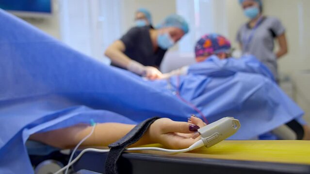 Heart rate sensor on the hand of female patient at surgical operation. Group of doctors working at backdrop in blur.