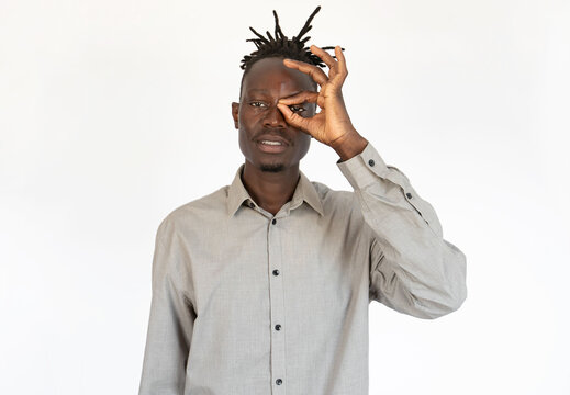 Portrait of confident young man peeking through fingers against white background. African American man with dreads wearing gray shirt looking at camera and smiling. Vision concept