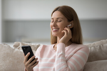 Serene woman wears earphones holds cell phone listen music through audio streaming and media services provider, use mobile application, spend carefree weekend at home using modern wireless technology