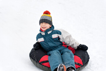Fototapeta na wymiar Happy boy in winter jumpsuit and hat with bubo rides on tubing in snow. Merry snowy winter fun