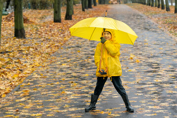 Smiling boy walks in the park with an yellow umbrella. Walk with child in rainy street