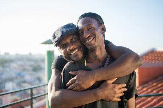 Close-up of happy African gay boyfriends looking at camera. Two bearded men in casual clothes standing together on roof top hugging taking photo on phone camera. LGBT peoples love, happiness concept