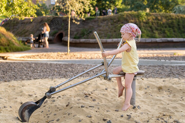 Little girl in bandana and summer clothes plays escalator simulator on playground in sandbox