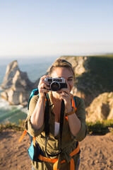 Portrait of mid adult woman traveling with camera. Happy female backpacker standing on top of mountain and taking photo. Travel and photographing concept