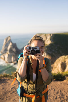 Portrait of mid adult woman traveling with camera. Happy female backpacker standing on top of mountain and taking photo. Travel and photographing concept