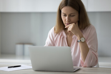 Millennial serious thoughtful female freelancer working on laptop seated in kitchen, staring at device screen looks busy makes on-line task, learn new software, having difficulties project or research