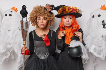 Shocked female magicians point at camera with embarrassed surprised expressions stand next to each...