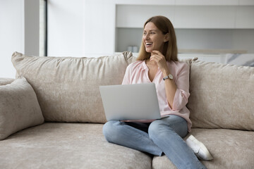 Dreamy woman staring into distance relaxing on cozy sofa with wireless computer on laps, enjoy modern technology usage for work or leisure, buying commercial services or communicates remotely at home