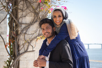 Portrait of young loving couple standing together gently holding hands. Young Muslim woman hugging from back happy smiling bearded man in park near sea. Relationship and romance concept