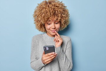 Curious cheerful woman with curly hair keeps index finger near lips focused at smartphone checks...