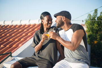 Fototapeta premium Portrait of African gay couple expressing love. Two bearded men dating on roof top sitting close smiling holding glasses of juice looking at each other with tender. LGBT couples love and life concept