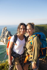 Portrait of smiling mother and daughter hiking together in mountains. Mid adult woman and teen girl posing against seascape. Active family weekend concept