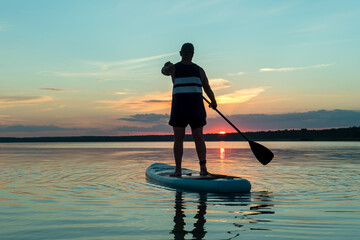 A man in shorts and a T-shirt on a SUP board with an oar against the backdrop of the sunset sky swim in the lake.