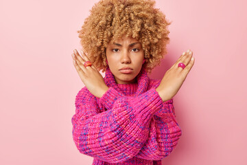 Irritated curly haired young woman crosses arms shows taboo gesture declines something rejects offer wears knitted jumper isolated over pink background prohibits something. Body language concept