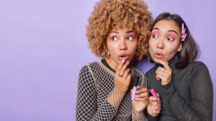 Horizontal shot of two ethnic female friends apply makeup hold lip gloss prepare for date wear black jumpers look amazed isolated over purple background empty space for your promotional content - obrazy, fototapety, plakaty