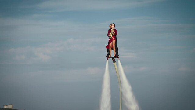 Show over the water on a flyboard. A woman in a red dress plays the saxophone, a man flies with her together.