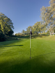 Obraz premium Golf Course green with blue sky and flag in view, autumn day with dew