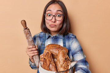 Horizontal shot of surprised Asian housewife purses lips holds rolling pin and homemade freshly baked bread wears transparent eyeglasses and blue shirt isolated over beige background. Bakery food