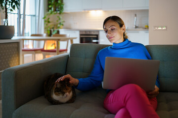 Happy young woman wearing blue sweater works on laptop and types important information for business project. Smiling businesswoman strokes domestic cat head and enjoys playing with pet in apartment