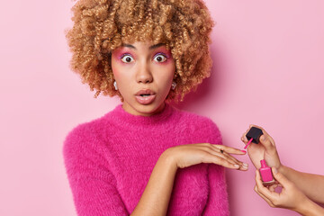 Amazed speechless curly woman with bright vivid makeup makes manicure applies nnail polish looks...