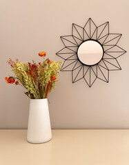 White vase with dried flowers in warm tones on a table, with a metal figure in the shape of the sun...