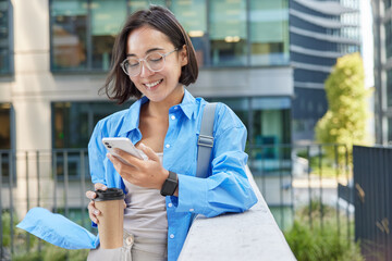 Outdoor shot of positive Asian woman holds cellular gadget and takeaway coffee checks received notification wears spectacles and blue shirt poses against urban background creats social publication