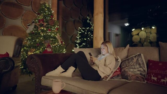 Full shot - Slow motion of a woman relaxing and laying on a sofa with Christmas pattern pillows, next to a decorated Christmas tree, looking and scrolling on her phone, on a calm wintery evening.
