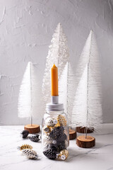 Fototapeta na wymiar Background with Christmas decorations. Glass candleholder with gold, black, silver cones and yellow candle, white holiday trees on grey textured background. Scandinavian minimalistic style. 