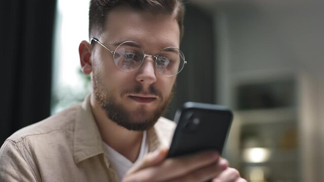 Young caucasian male in round eyeglasses scrolling smartphone, checking social media and e-mail.