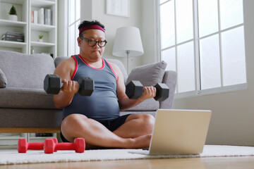 Fat middle-aged asian man in sportswear using dumbbell exercising following trainer online via computer laptop at home - Powered by Adobe