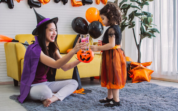 Asian beautiful woman or mother and little Caucasian sweet girl wearing witch costume with hats, playing trick or treat with fun, smiling with happiness, celebrating Halloween party at home.