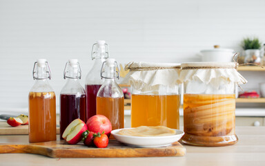 Portrait glass bottles of Kombucha healthy fermented probiotic tea drinks with ingredients red apples, strawberries, mushroom scoby on wooden table in comfortable kitchen at home. Beverange Concept.