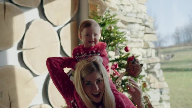 Mid shot - Smiling baby sitting on mom's shoulders wearing matching Christmas pyjama set and dancing in front of a Christmas tree in a living room 