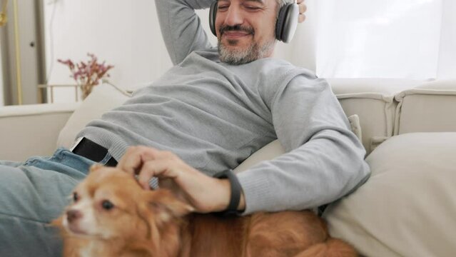 Handsome Senior mature adult man sitting on the couch listening to music with headphones and playing with chihuahua dog, Lifestyle concept, Relax in living room at home.