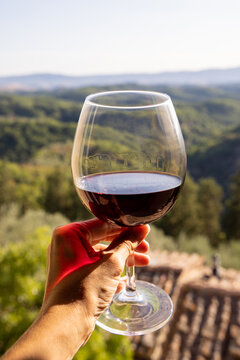 Woman holding a glass of red wine with beautiful landscape of Italy in a background on a sunny day. View from the window.
