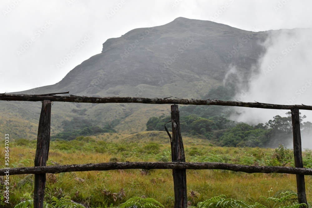 Poster scenic view of a hill covered in fog and clouds with a wooden fence and green slope in front
