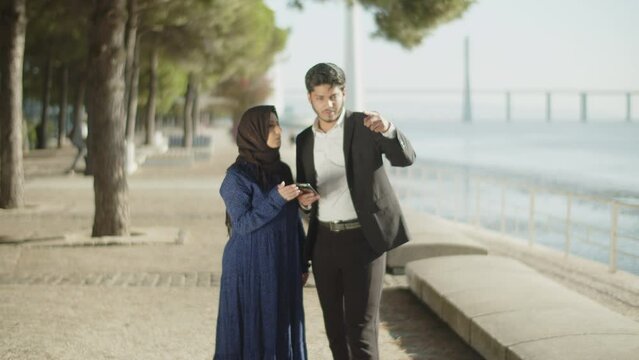 Arabic couple from Middle East using smartphone for destination, dark-haired man holding phone and showing direction to his wife, woman looking at phone and helping him. Travelling, location concept