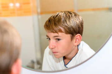 Preteen looks at red face with acne in the bathroom mirror with serious expression
