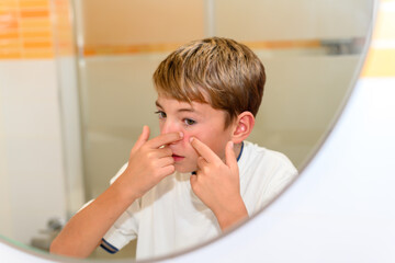 A preteen touches a pimple with his fingers in the toilet mirror.