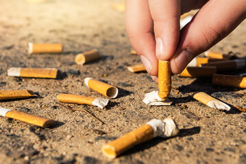 A hand puts out a cigarette on the ground littered with cigarette butts. Smoking harms a person's health. Cigarettes