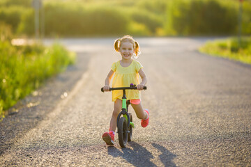 Happy beautiful little girl fast running and riding on first bike without pedals on road at town in warm summer day. Cute 3 years old toddler. Front view. Learning to keep balance. Sunset light.
