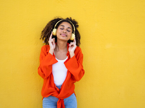 Young african american teenage girl listening to music happily with headphones over yellow wall