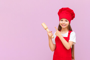 A little baker girl in a chef's hat and apron holds a rolling pin and points at your advertisement on a pink isolated background. Copy space.