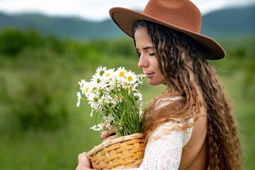 A middle-aged woman in a white dress and brown hat holds a lA middle-aged woman in a white dress and brown hat holds a basket in her hands with a large bouquet of daisies.arge bouquet of daisies in