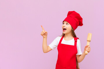 A little baker girl in a chef's hat and apron holds a rolling pin and points at your advertisement on a pink isolated background. Copy space.