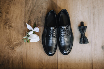 The groom's black shoes, bow tie, boutonniere lie on a wooden background.Wedding photo of...